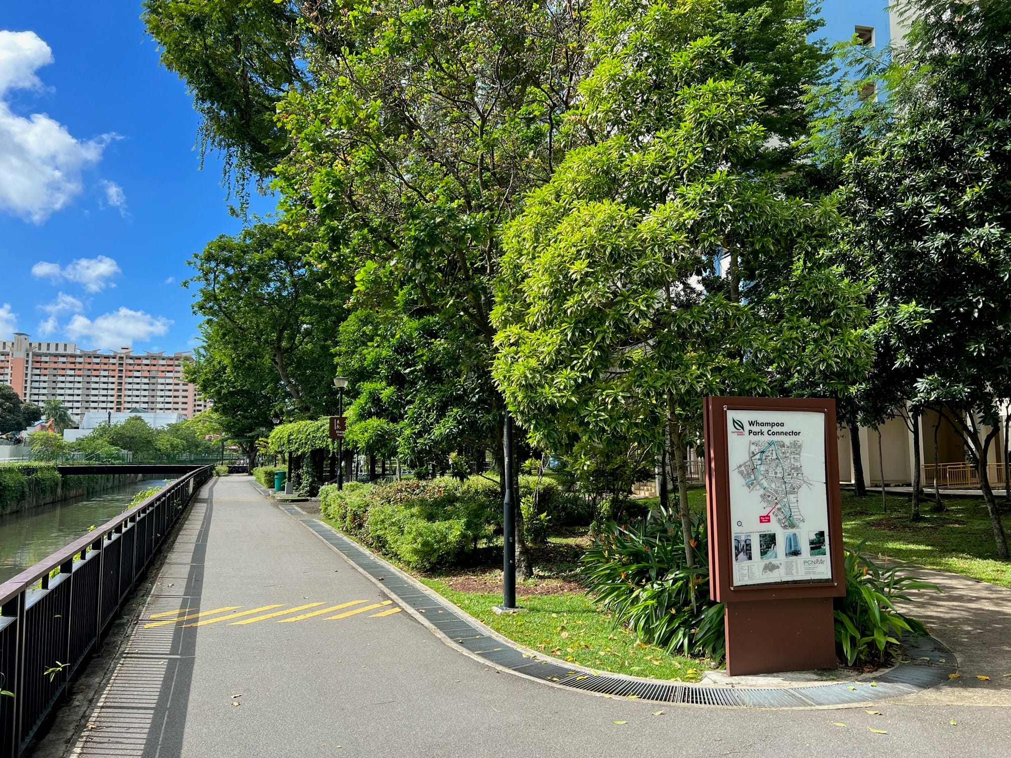 Pathway near a canal lined with lush trees, a park connector map sign, and buildings in the background under a blue sky.
